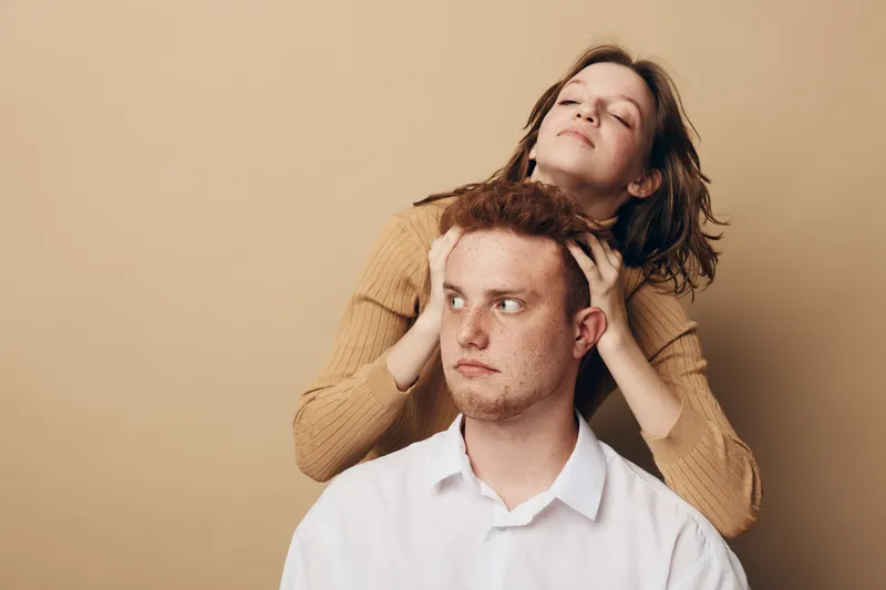 Young woman with closed eyes massages head of serious young man in white shirt on beige background. Concept of relaxation, stress relief, and emotional support.