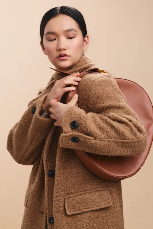 asian woman wearing a warm brown coat with a round leather bag stands against a soft beige backdrop, exuding calm confidence, elegance, and modern poise.