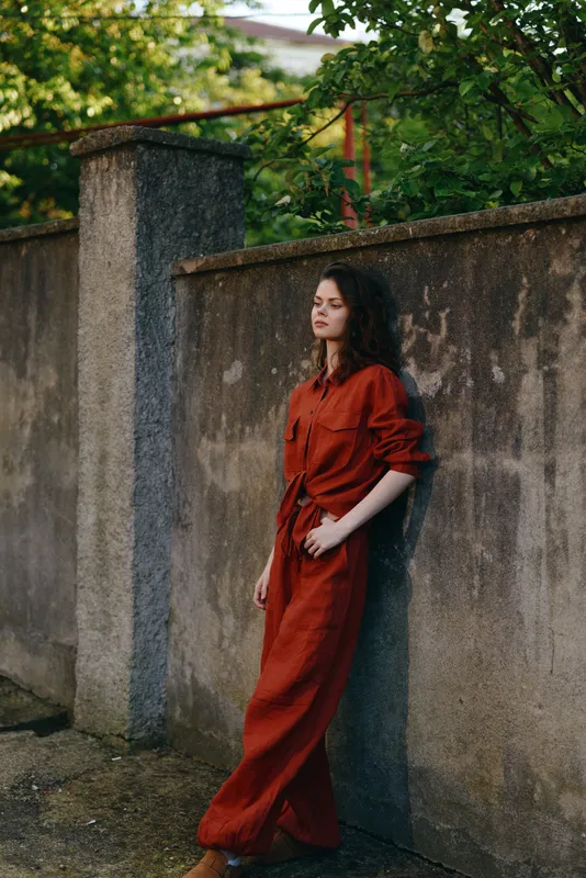 Young woman in a red dress stands against a weathered wall outdoors, relaxed pose, natural light, contemplative mood, lush greenery framing the scene, gentle breeze, timeless atmosphere