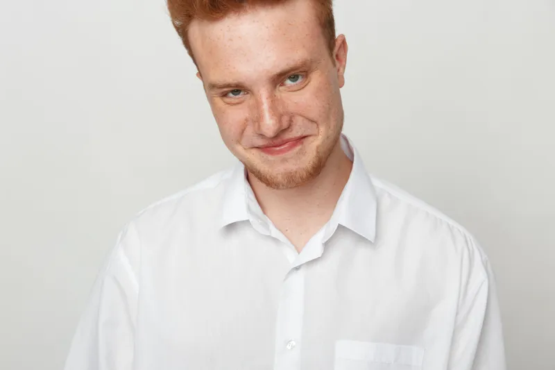Smiling young man with red hair and beard wearing white shirt looks at camera with friendly expression on light background. Concept of casual portrait, happiness, and confidence.