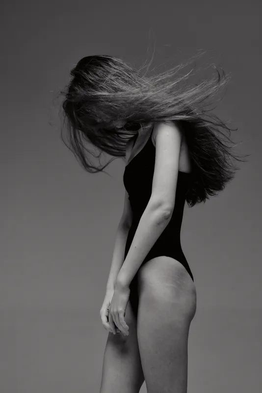 Black and white portrait of a young woman with long hair in motion wearing a black bodysuit.