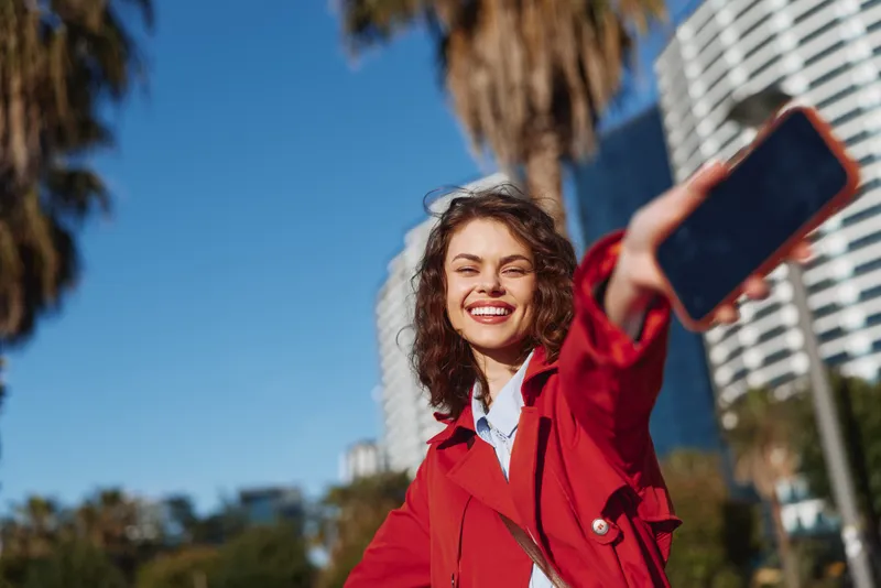 A young woman in a red coat takes a selfie on a sunny city street, smiling as she holds a smartphone toward the camera, with palm trees in the background today.