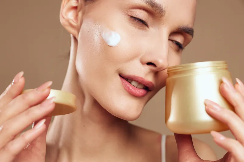 A young woman applies face cream on her cheek using her fingers while holding a jar of moisturizer in a close-up beauty skincare routine. Concept for skin hydration and self-care.