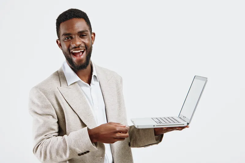 Joyful young black man in beige blazer holding open laptop, smiling broadly while looking at camera against plain white background, casual professional portrait, happy expression, technology, office l
