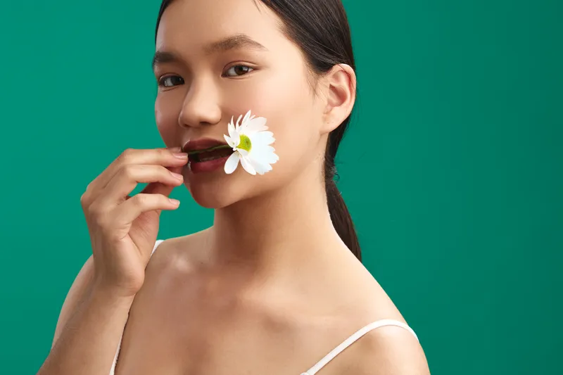 Asian woman with a flower in her mouth stands against a vivid green backdrop, creating a minimalist portrait that exudes calm, poise, and modern beauty.