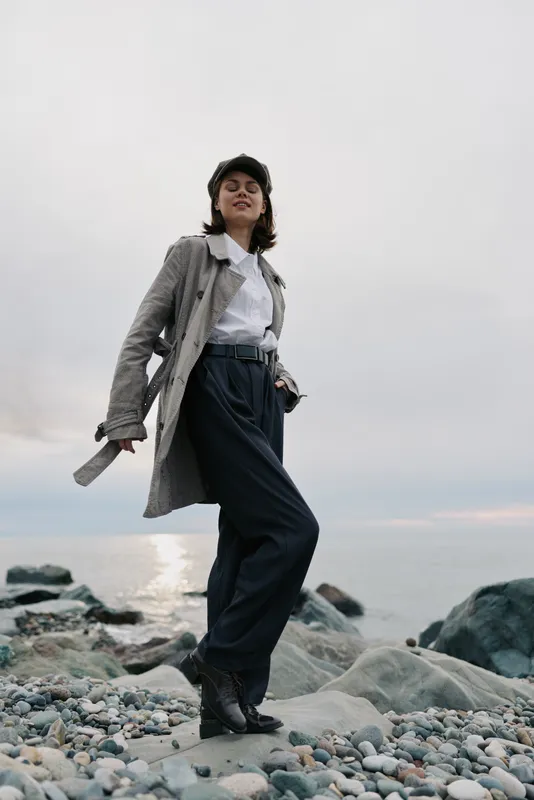 Young woman in trench coat and high waist pants stands confidently on rocky beach near sea under cloudy sky. Concept of fashion, confidence, and outdoor lifestyle with natural background.