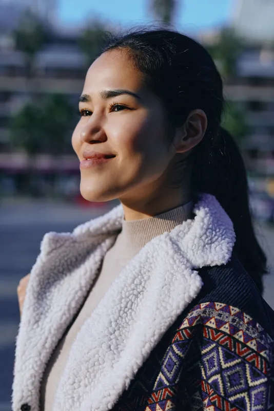 Woman in a cozy shearling coat stands outdoors, sunlight on her face and a calm smile, urban backdrop blurred, fashion and warmth captured in a natural profile shot