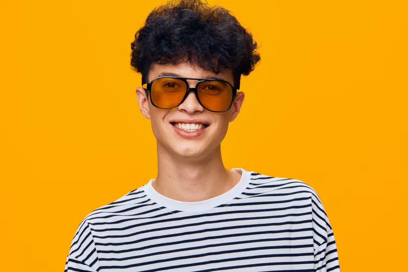 smiling man with curly hair wearing brown sunglasses and striped shirt stands against bright orange background, showcasing casual style and youthful vibe.