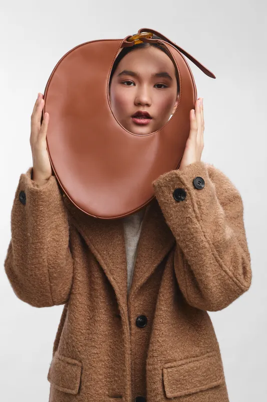 Asian woman wears a stylish wool coat and a large circular brown accessory framing her face in a studio, creating a chic fashion portrait with a minimal background and confident mood.
