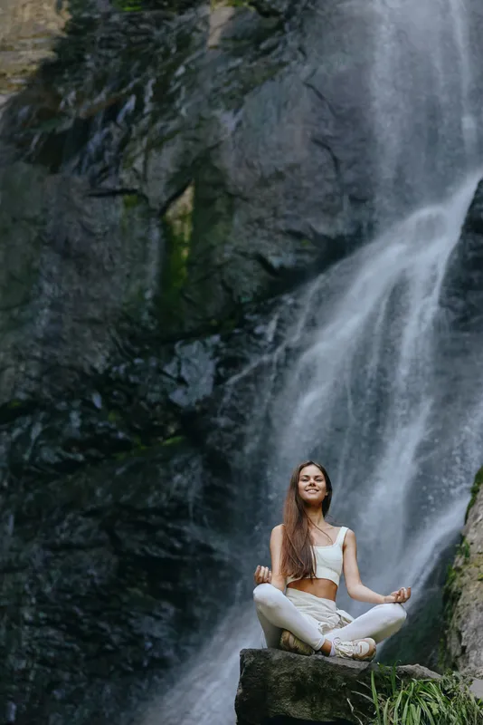 Young woman meditating peacefully near waterfall, sitting cross-legged on rock with serene expression surrounded by lush nature and flowing water, in casual white clothes.