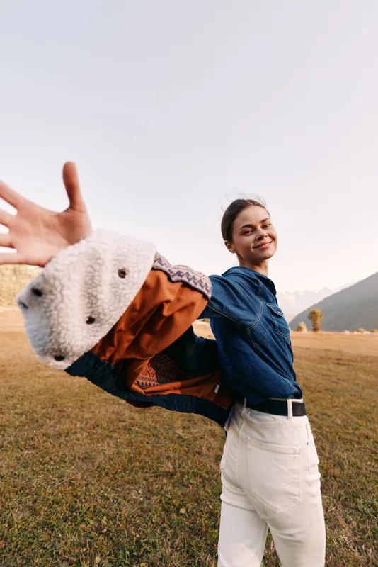Woman smile outdoors in a field with jacket thrown back, playful young portrait by mountains, casual denim and white pants, sunlight and carefree motion capturing joyful lifestyle moment.