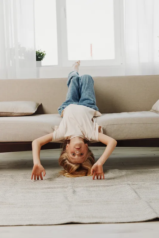 Playful child in casual clothes hangs upside down on beige sofa in bright living room. Concept of childhood fun, home leisure, and carefree moments with natural light and relaxed atmosphere.