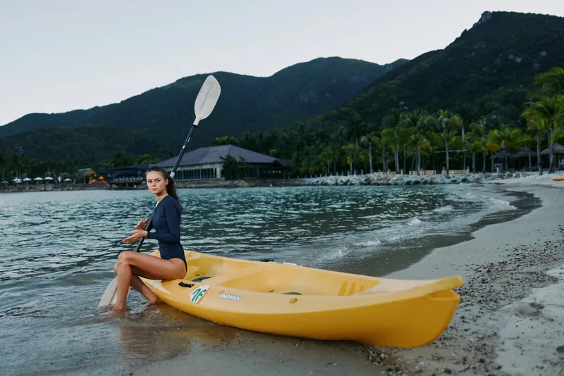 Woman kayaking on calm beach shore at dusk in tropical location with mountains and palm trees in background, enjoying outdoor water sports and adventure.