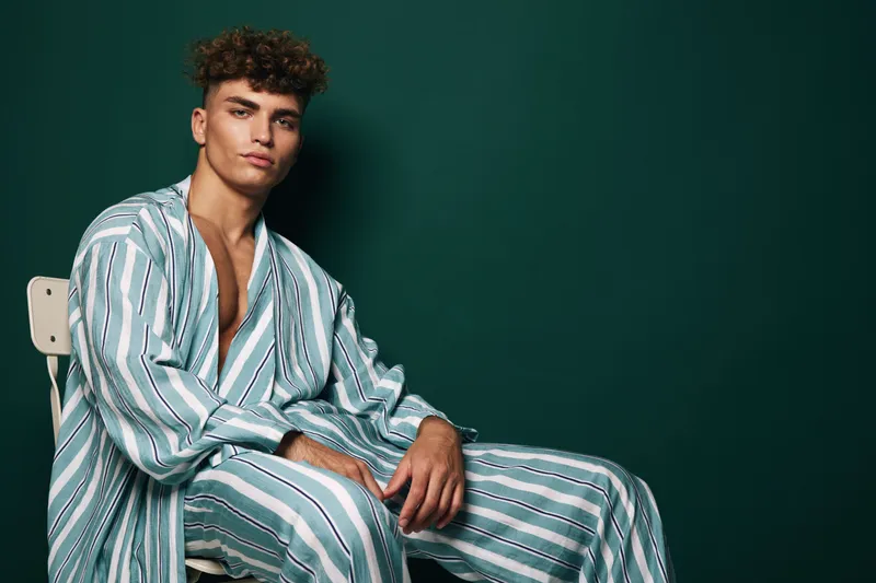 Young man with curly hair wearing striped pajamas sitting on chair against dark green background in studio portrait with relaxed pose and confident expression.