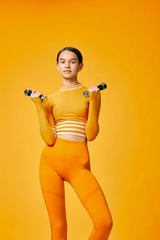 Fitness woman in bright orange activewear holding dumbbells in a studio with bold orange backdrop, showcasing strength, balance and focus during a workout session.