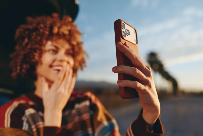 Woman with curly hair smiling, smartphone selfie on a street at sunset, close up, shallow depth of field, blurred outdoor background, warm golden light, lifestyle moment, friendly portrait