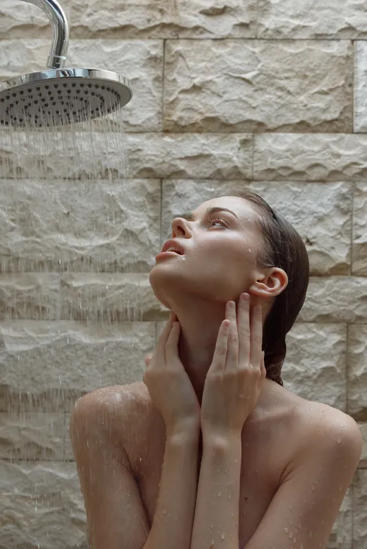 young woman enjoying relaxing warm shower under rainfall showerhead against beige textured wall, peaceful moment of self-care and freshness