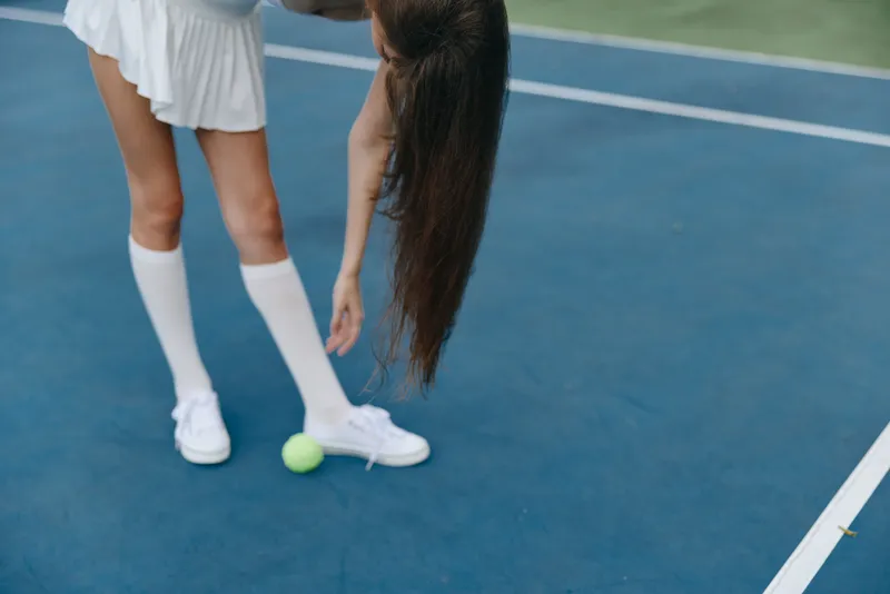 Young female tennis player in a white outfit reaching for a tennis ball on a blue court, showcasing athleticism and focus