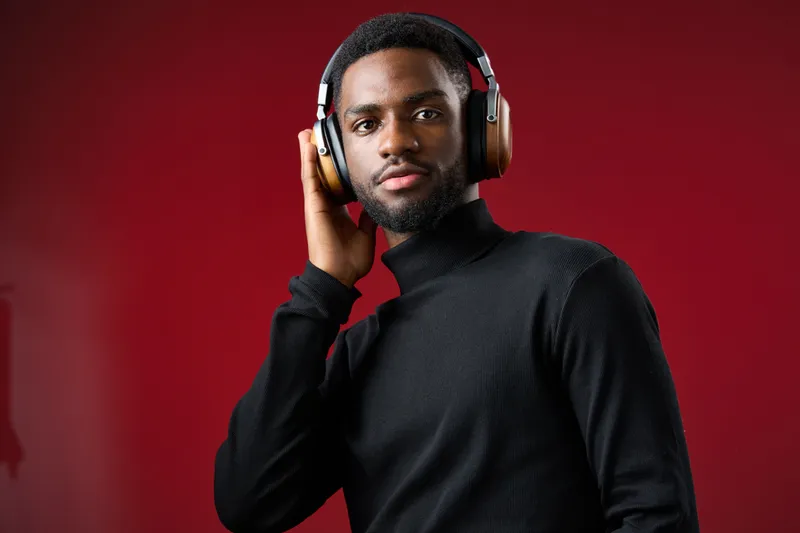 Young African man enjoying music with stylish headphones against a vibrant red background, conveying emotions of relaxation and focus while dressed in a chic black turtleneck