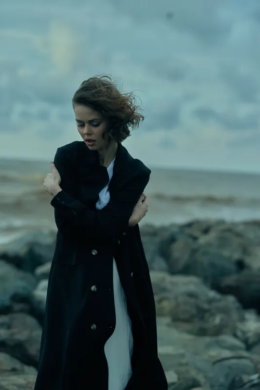 Windy coastline scene featuring a woman in a long dark coat, hair blown by gusts, standing on rocky shore with a moody sea backdrop, dramatic fashion mood and stormy atmosphere, isolated