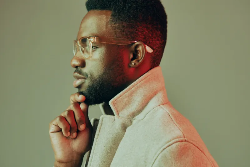 Thoughtful young man in stylish gray coat with glasses, posing against a soft green background, reflecting confidence and contemplation