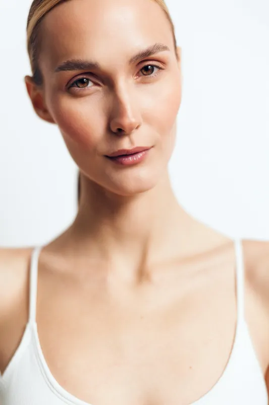 Portrait of young woman with natural skin and minimal makeup wearing white tank top looking confidently at camera against white background in studio light.