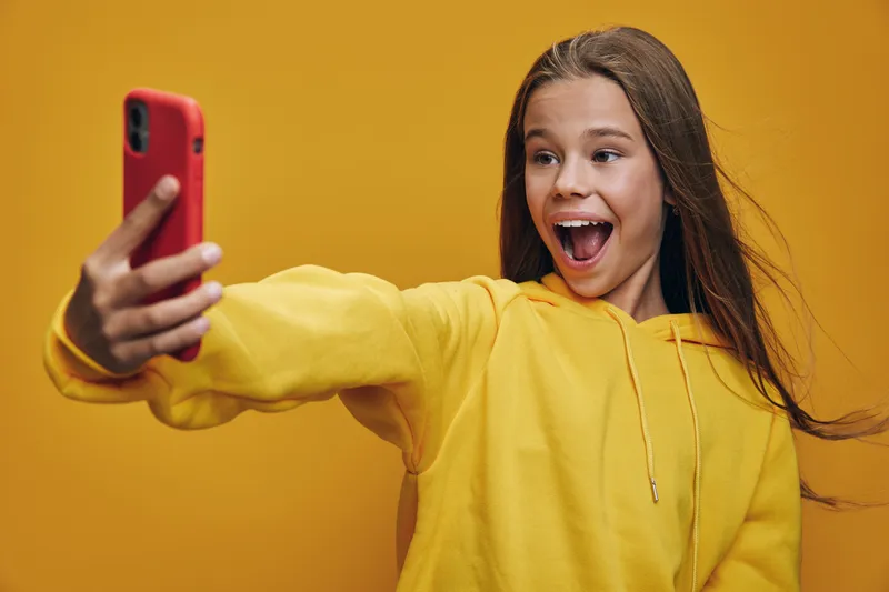 Girl wearing yellow hoodie taking selfie with smartphone on yellow background, smiling and excited, capturing fun moment with long hair flowing to the side.