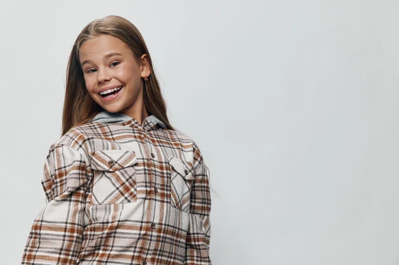 Girl smiling and posing in plaid shirt with long hair on gray background. Happy child portrait with casual fashion for lifestyle, youth and positive expression indoors.
