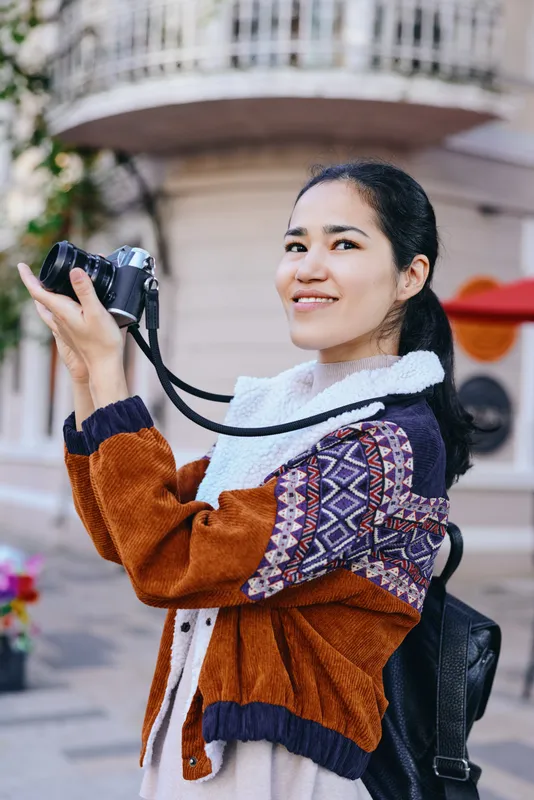 Young woman with camera on street smiles while shooting, wearing a cozy knit cardigan and backpack, urban backdrop, candid outdoor moment for travel, fashion and lifestyle photography