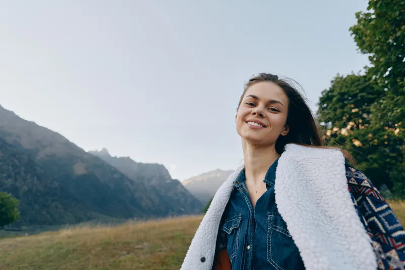 Woman smiling portrait in mountains and nature outdoors, happy traveler wearing denim and shearling jacket, enjoying scenic field landscape, relaxation and adventure in fresh air.