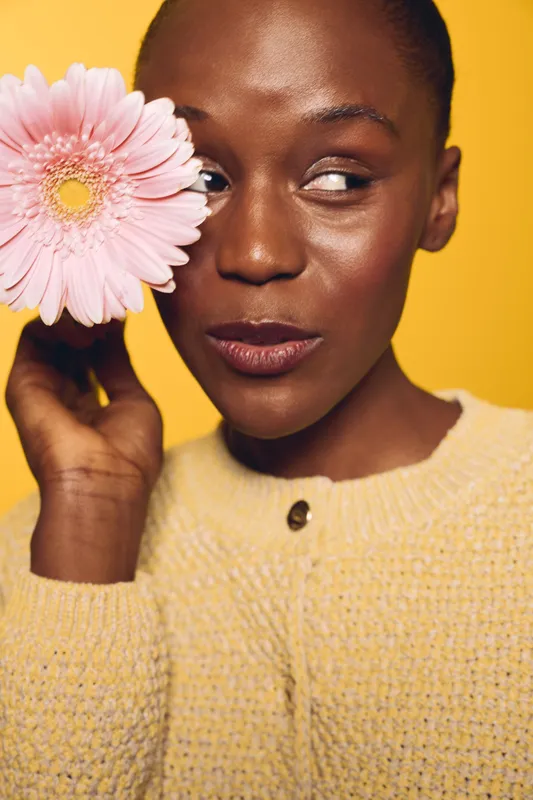 Close-up of a dark skinned African woman holding a pink flower near her eye. She wears a yellow sweater and looks sideways on a bright yellow background. Concept of beauty and nature.