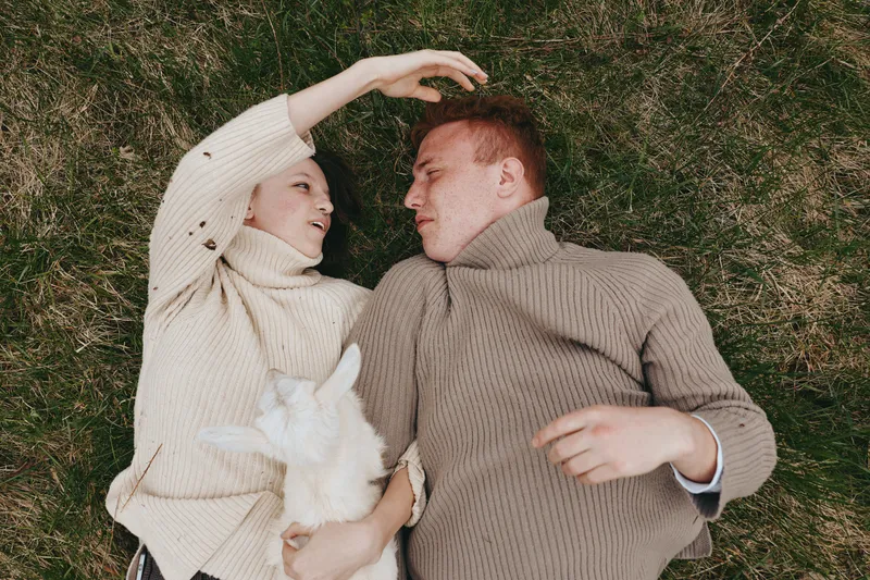 Young couple lies on grass wearing warm sweaters while holding a small white goat, enjoying a relaxed lifestyle moment in nature with affectionate and calm expressions.