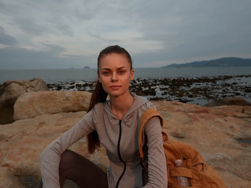 Young woman sitting on a rocky beach at sunset, wearing a grey athletic outfit with a backpack, looking contemplative and serene amidst the natural backdrop