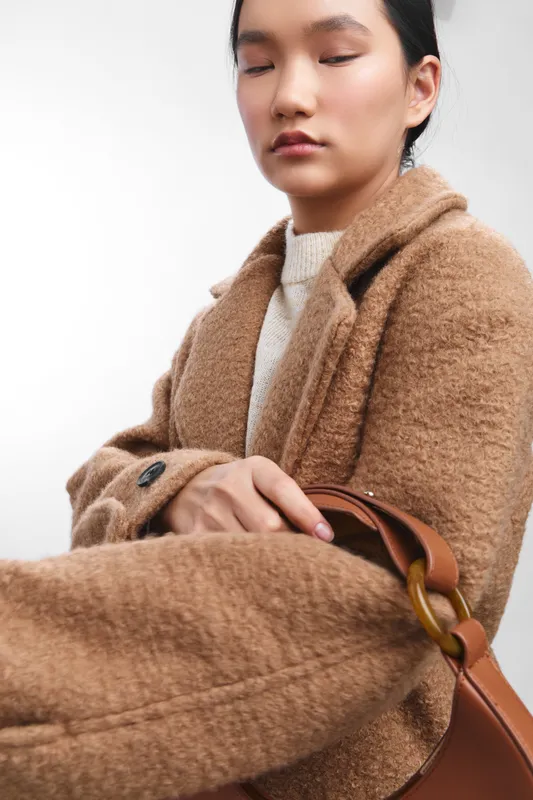Asian woman wearing a tan coat rests with a calm expression, eyes closed, exuding elegance and focus in a clean minimalist studio setting.