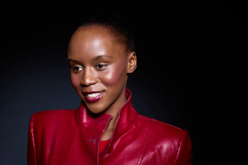 Portrait of a smiling black woman with dark skin wearing a red leather jacket on black background. Studio lighting highlights her natural beauty and confident expression.