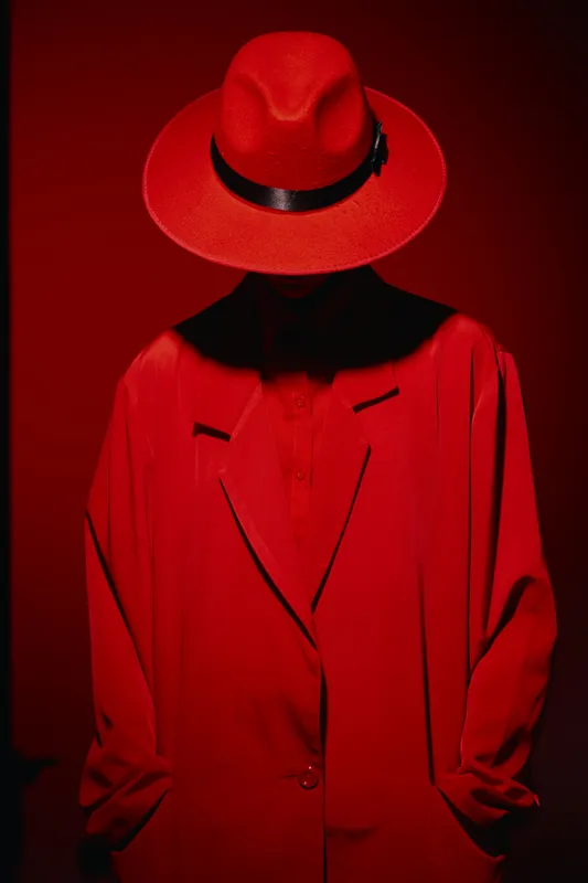 Mysterious figure in a red hat and oversized red jacket stands against red backdrop, face obscured by shadow. Concept for fashion, beauty, and creative style.