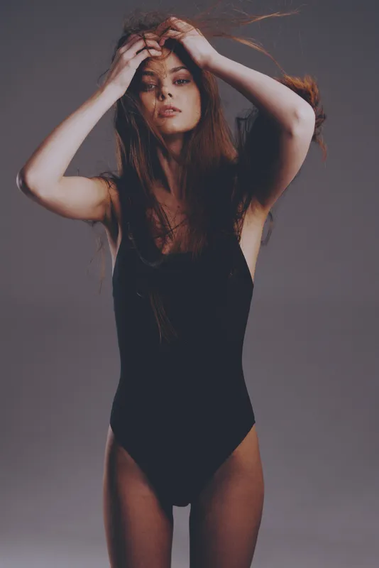 slender woman in black bodysuit posing with hands in hair against gray background