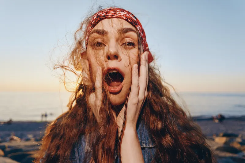 Lifestyle woman with boho-western style wearing denim jacket and red bandana, expressing surprise in street style with film color tones at sunset beach.