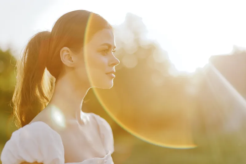 Young woman in a white dress with a ponytail, enjoying a sunny day, showcasing a serene expression, golden hour lighting, nature background, summer vibes