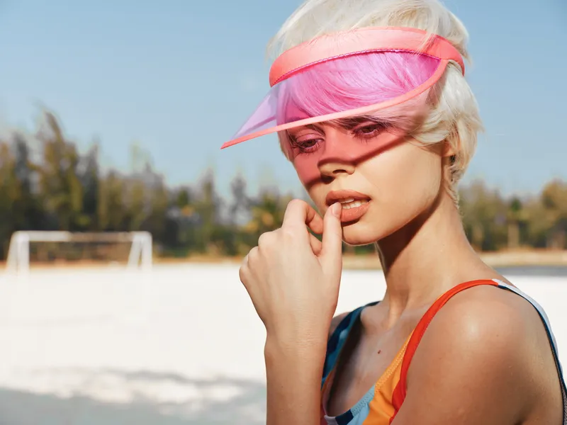 Portrait of a stylish young woman wearing a bright pink visor, sunlit skin, and bold beachwear, captured in a warm outdoor setting with distant sports elements.