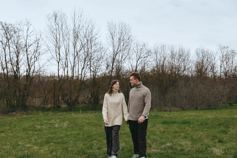 Cheerful couple outdoors, walking hand in hand through a sunny field, cozy knitwear, soft breeze, natural colors, bare trees in the background, relaxed mood, genuine connection, daytime happiness