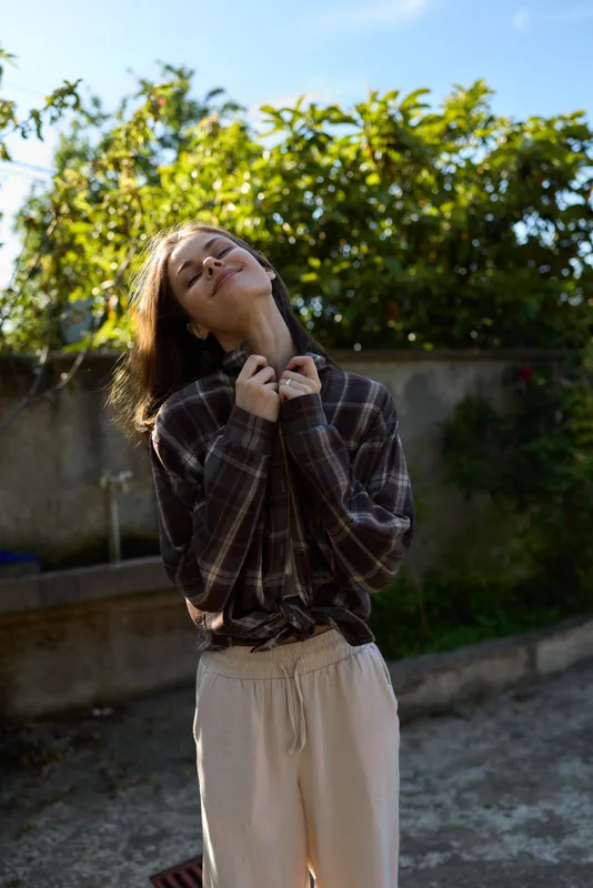 Casual lifestyle portrait of a woman enjoying a sunny outdoor moment, wearing plaid shirt and light pants, tilting her head with a relaxed smile, natural garden backdrop