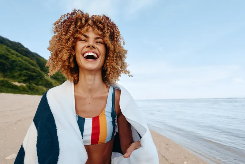 Smiling woman with curly hair walking on a sandy beach by the ocean, wearing a swimsuit and denim jacket, bright summer daylight, carefree vacation mood, travel lifestyle, outdoor wellness and freedom