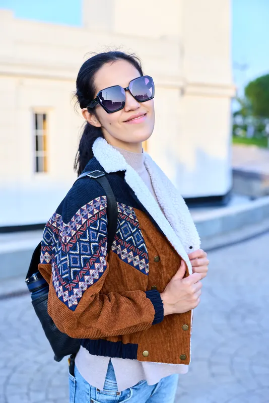 Fashionable street style of a woman with a patterned brown jacket, jeans, scarf and backpack, posing outdoors with sunglasses under a sunny sky, displaying casual confidence and contemporary urban