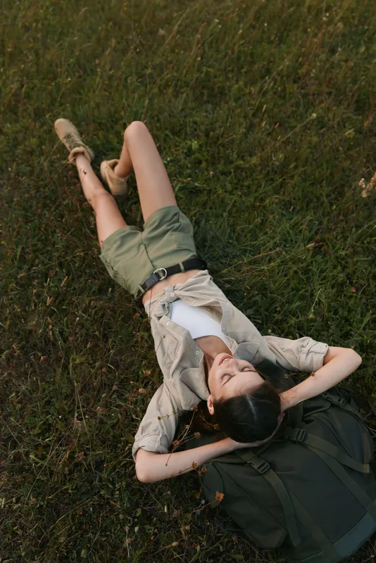 Woman relaxing outdoors lying on grass with backpack nearby, casual travel outfit, captured from above, lifestyle moment in nature
