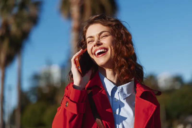 Young woman with curly hair laughs happily while talking on a smartphone outdoors in a red coat under clear blue sky with blurred trees in the background.