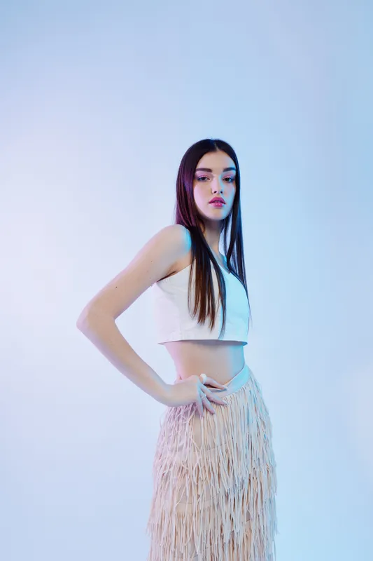 Young woman in a white crop top and fringe skirt poses confidently against a soft blue background. Concept of beauty, fashion, and modern style.
