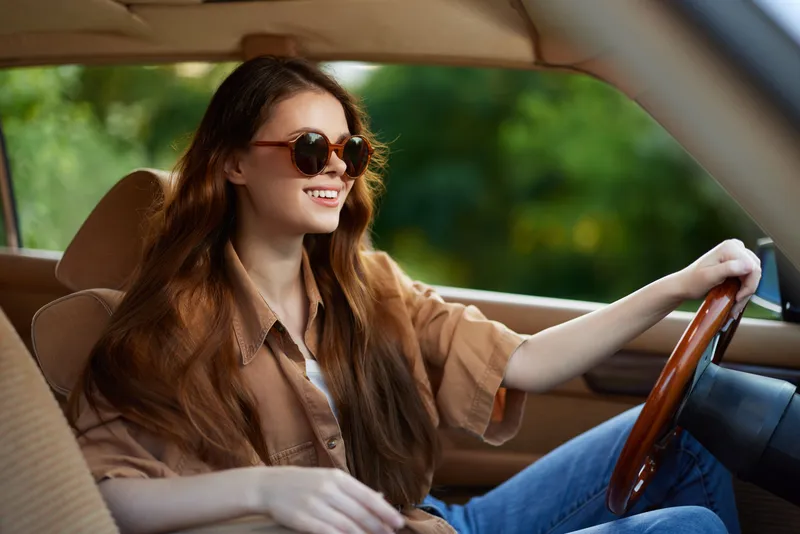 Happy young woman driving a car with sunglasses, enjoying a sunny day, displaying a casual style and vibrant nature background.