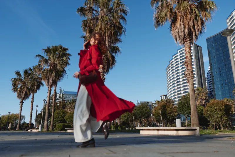 A fashionable woman in a flowing red coat strides along a sunny city street, with palm trees and modern towers in the background, conveying motion and urban style.