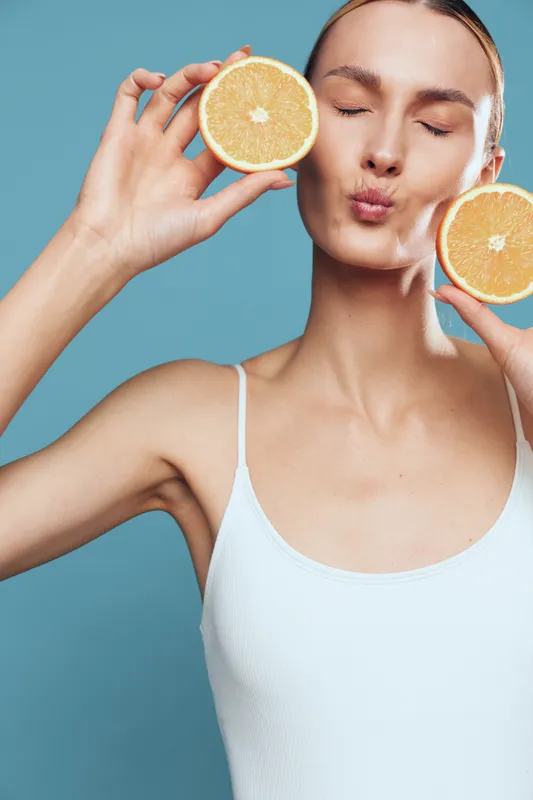 A young woman in a white tank top holds two halves of a fresh orange near her face with closed eyes and puckered lips. The blue background highlights the healthy lifestyle concept.